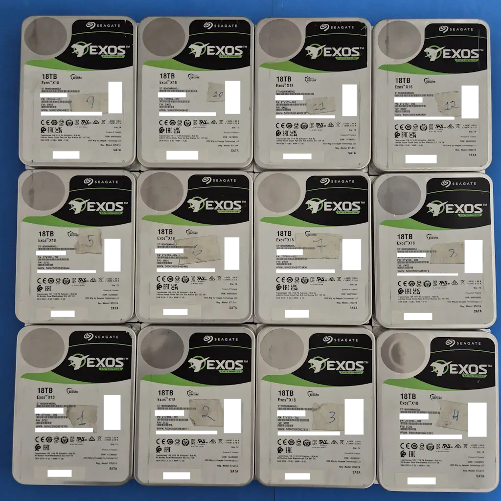 Sixteen Seagate Exos 18TB hard drives arranged in a four-by-four grid on a blue surface, showing white enclosures with green/black labels.